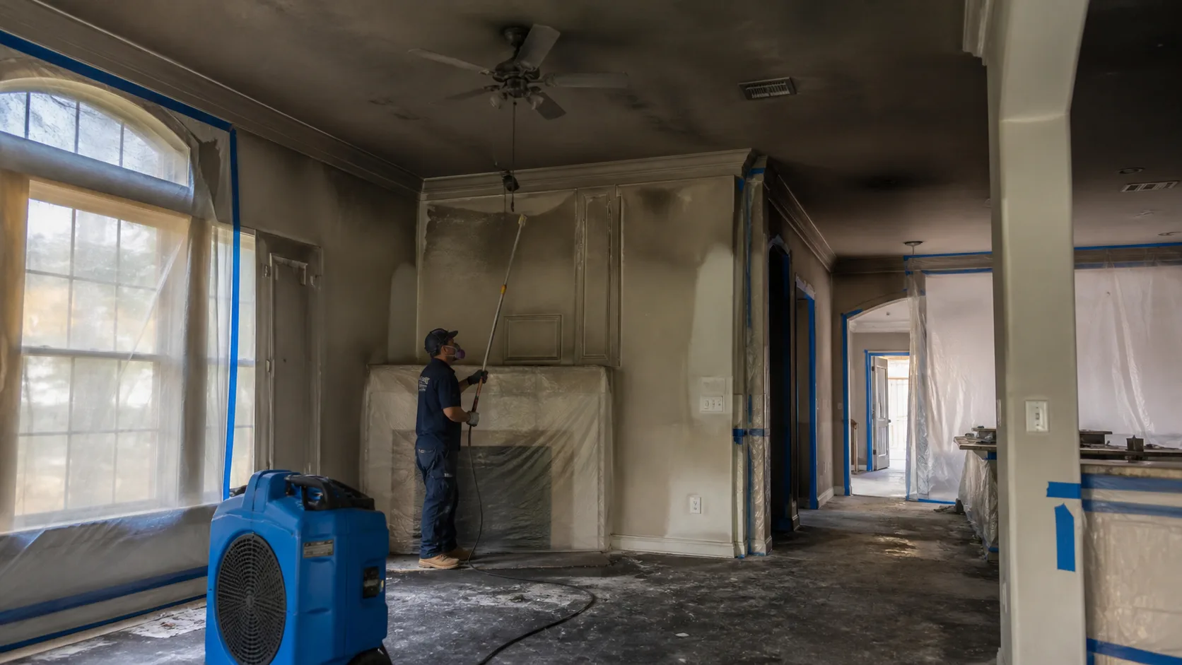 A technician in a respirator mask and work uniform uses a long roller to paint a wall in a room with a dark, smoke-damaged ceiling, covered windows, and a blue air mover on the floor.