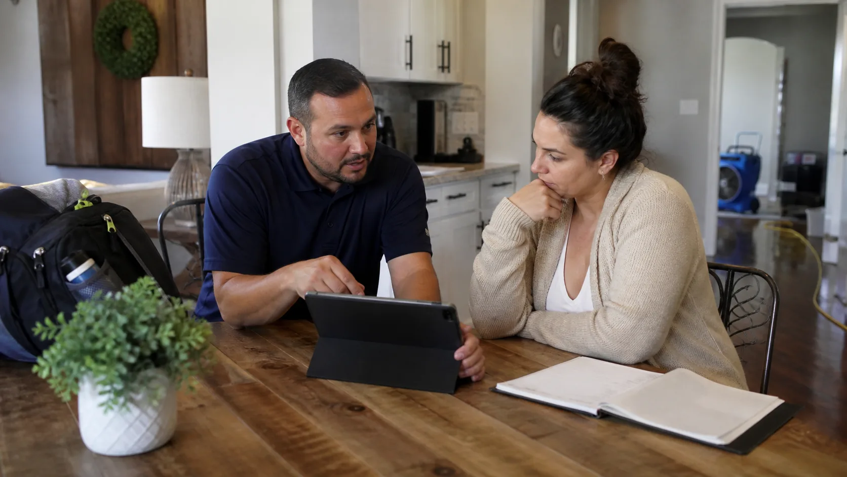 A homeowner and an adjuster sit at a wooden table, with the adjuster pointing at a tablet and the homeowner listening intently, while water damage restoration equipment is visible in the background.