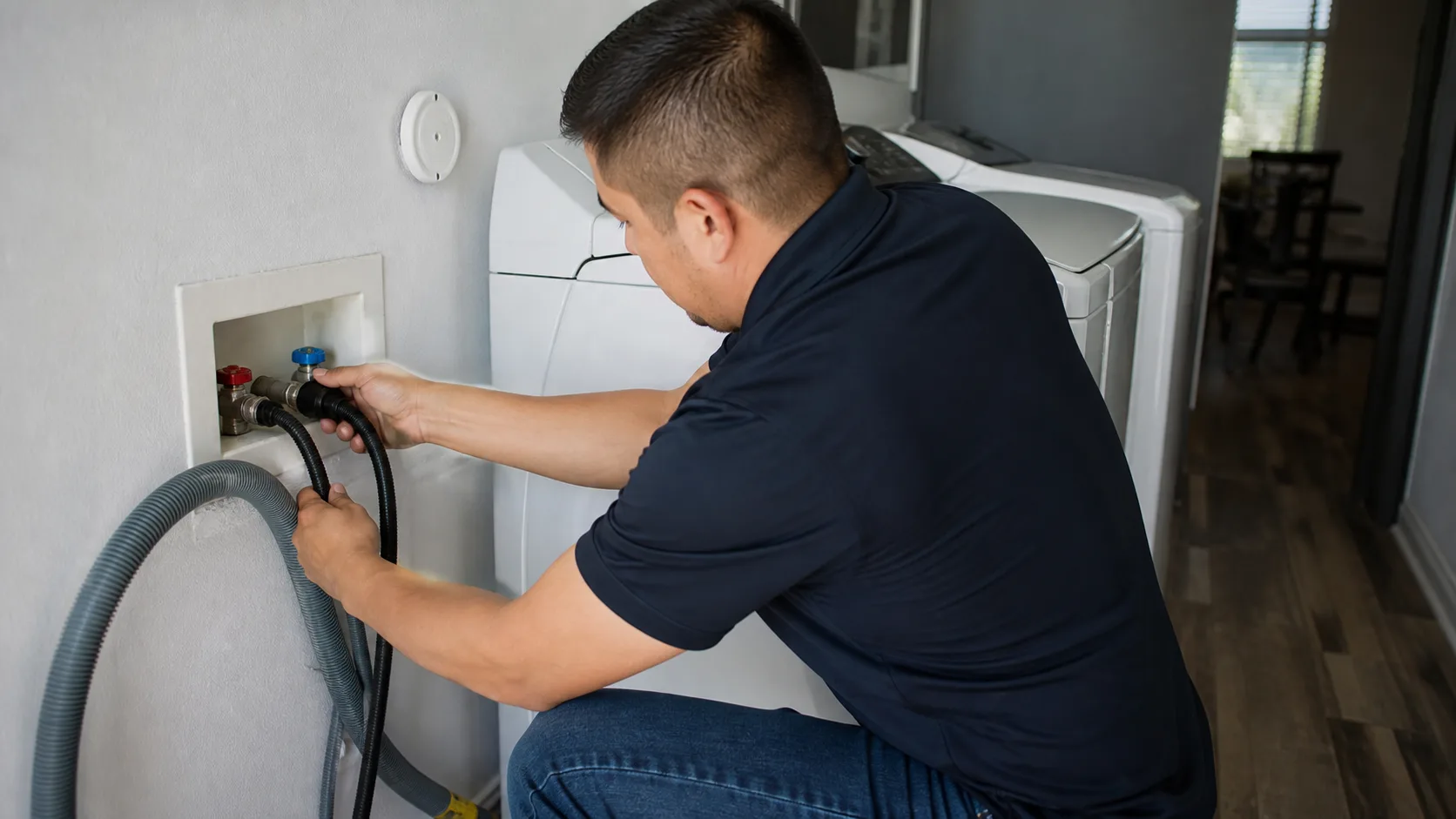 A technician wearing a dark blue shirt and jeans crouches to connect hoses to a washing machine in a laundry room.