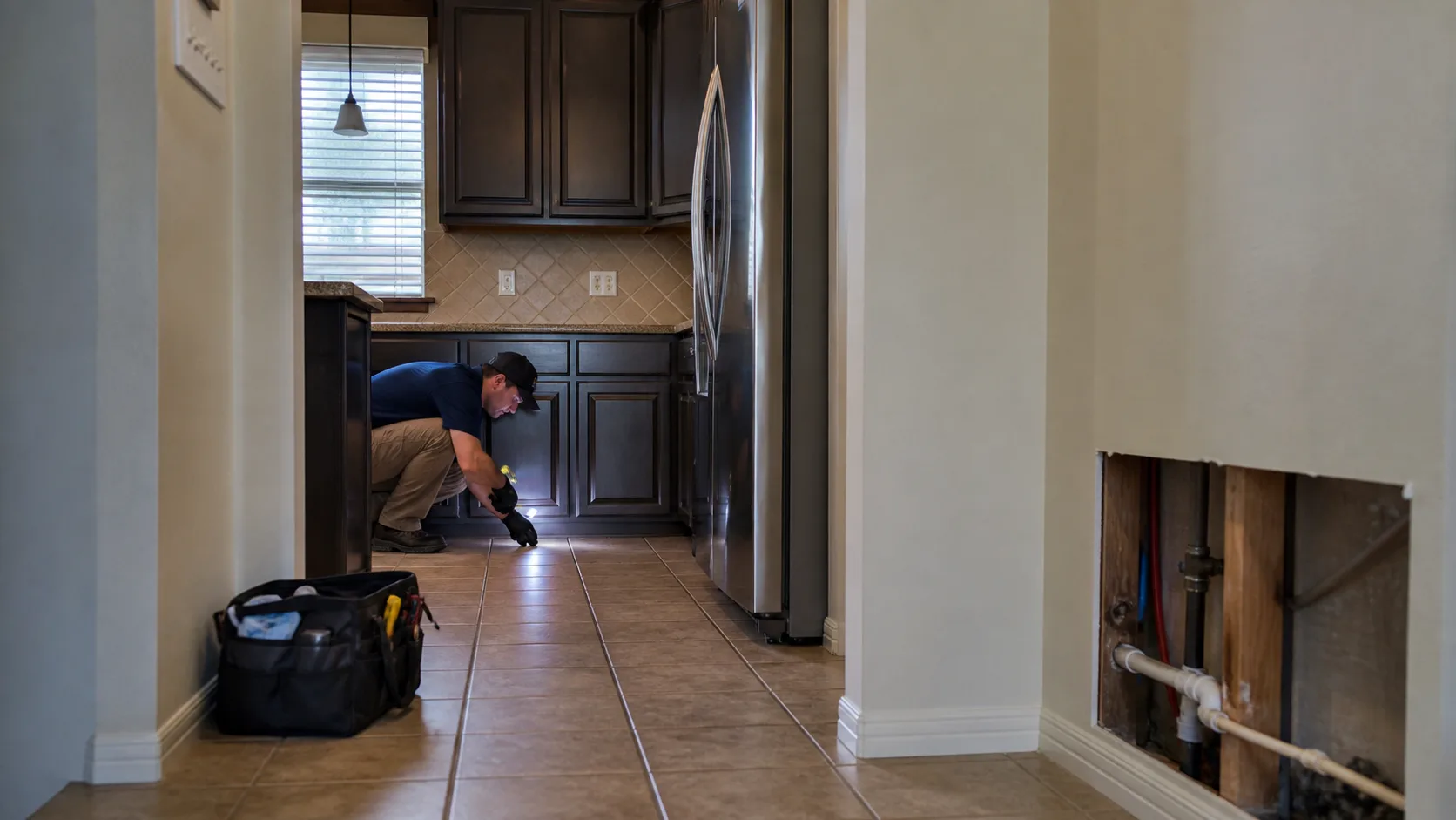 A technician wearing a cap and gloves crouches in a kitchen, shining a flashlight under dark cabinets, with a toolbox nearby and an exposed wall with pipes in the foreground.